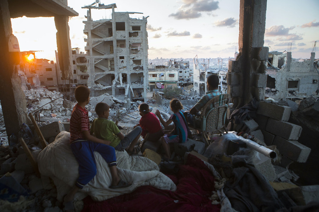 一家巴勒斯坦人坐在他们位于加沙塔法（At-Tuffah）区、被以色利空袭严重摧毁的房子上，2014年9月21日. 在持续7周的袭击中， 有2131名巴勒斯坦人丧生，其中包括501名儿童，估计有18,000间房屋遭到摧毁或严重破坏，令108,00人无家可归. 摄影：安·帕克.